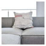 Funda con cojín cuadrada Antología color blanco y rosa con tejido artesanal decorativo, ubicada sobre sofá gris en ambiente minimalista, foto plano americano. All Star muebles