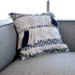 Funda con cojín cuadrada Antología color blanco y azul con tejido artesanal decorativo, ubicada sobre sofá gris, foto plano detalle. All Star muebles