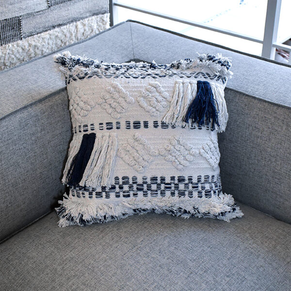 Funda con cojín cuadrada Antología color blanco y azul con tejido artesanal decorativo, ubicada sobre sofá gris en ambiente minimalista, foto plano americano. All Star muebles