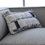 Funda con cojín rectangular Antología color blanco y azul con tejido artesanal decorativo, ubicada sobre sofá gris, foto plano detalle. All Star muebles