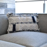 Funda con cojín rectangular Antología color blanco y azul con tejido artesanal decorativo, ubicada sobre sofá gris en ambiente minimalista, foto plano americano. All Star muebles