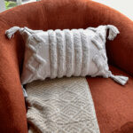 Funda con cojín rectangular Rustic color blanco con textura afelpada, ubicada sobre sofá rojo con una manta blanca complementaria, foto plano americano. All Star muebles