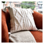 Funda con cojín Rustic color blanco con textura afelpada, ubicada sobre sofá rojo con una manta blanca complementaria, foto plano americano. All Star muebles