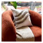 Funda con cojín Rustic color blanco con detalles avellana y textura afelpada, ubicada sobre sofá rojo con una manta blanca complementaria, foto plano americano. All Star muebles