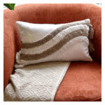 Funda con cojín rectangular Rustic color blanco con detalles avellana y textura afelpada, ubicada sobre sofá rojo con una manta blanca complementaria, foto plano americano. All Star muebles