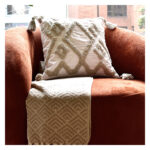 Funda con cojín Rustic color blanco con detalles beige y textura afelpada, ubicada sobre sofá rojo con una manta blanca complementaria, foto plano americano. All Star muebles