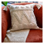 Funda con cojín Rustic color blanco con textura afelpada, ubicada sobre sofá rojo con una manta blanca complementaria, foto plano americano. All Star muebles