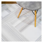 Fotografía plano detalle de tapete geométrico lineal en tonos grises con plateado, con una silla de patas en madera encima. All Star decoración.