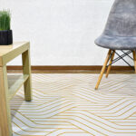 Ambiente de sala minimalista moderno con mesa auxiliar en madera, silla tapizada gris con patas en madera y tapete gris con textura geométrica dorada. All Star decoración