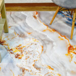 Fotografía plano detalle de tapete con textura mármol en tonos grises con plateado y naranja, con una silla de patas en madera encima. All Star decoración.