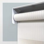 Detalle del mecanismo de una persiana enrollable tipo zebra con cadena plástica blanca y soporte lateral, textura de tela. All Star decoración