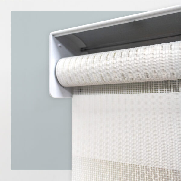 Detalle del mecanismo de una persiana enrollable tipo zebra con cadena plástica blanca y soporte lateral, textura de tela. All Star decoración