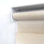 Detalle del mecanismo de una persiana enrollable tipo zebra con cadena plástica blanca y soporte lateral, textura enlazada vertical. All Star decoración