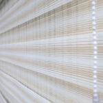 Detalle de persiana enrollable tipo zebra en tonos beige y blanco con textura de tela. All Star decoración