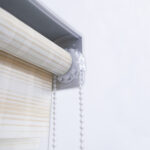 Detalle del mecanismo de una persiana enrollable tipo zebra con cadena plástica blanca y soporte lateral. All Star decoración